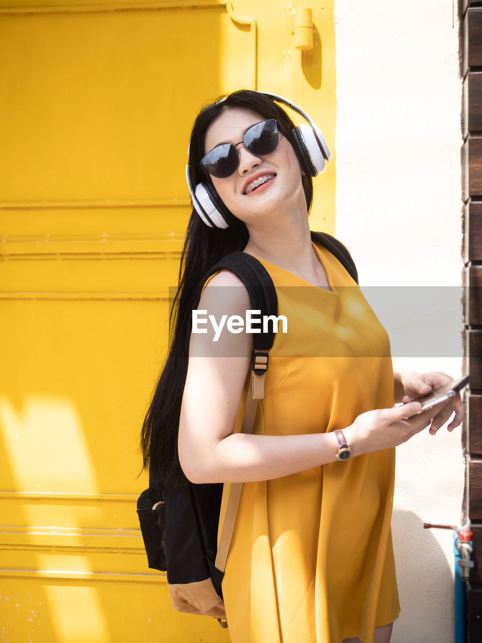 Smiling young woman holding mobile phone while standing against yellow door