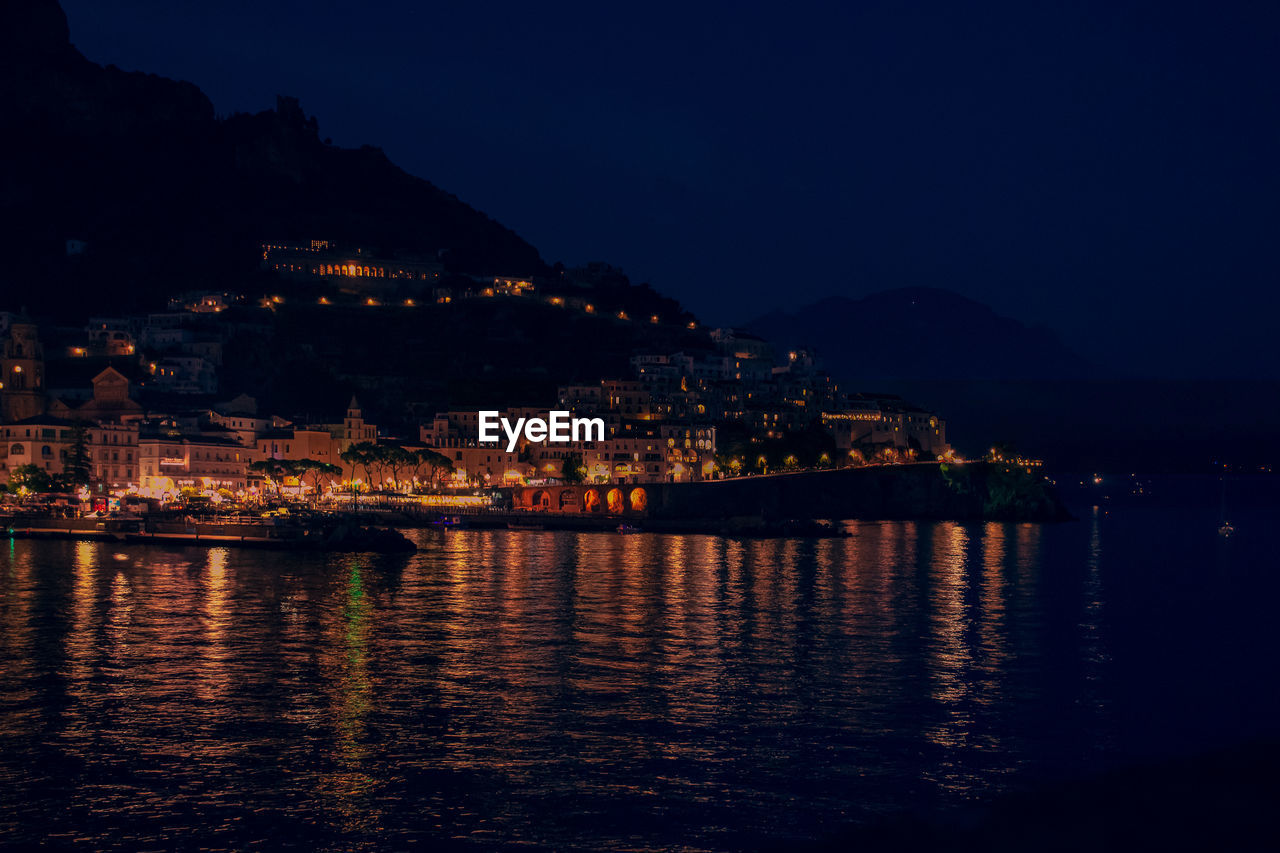 Illuminated buildings by river against sky at night