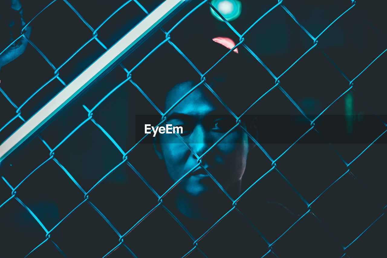 Close-up portrait of man seen through chainlink fence at night