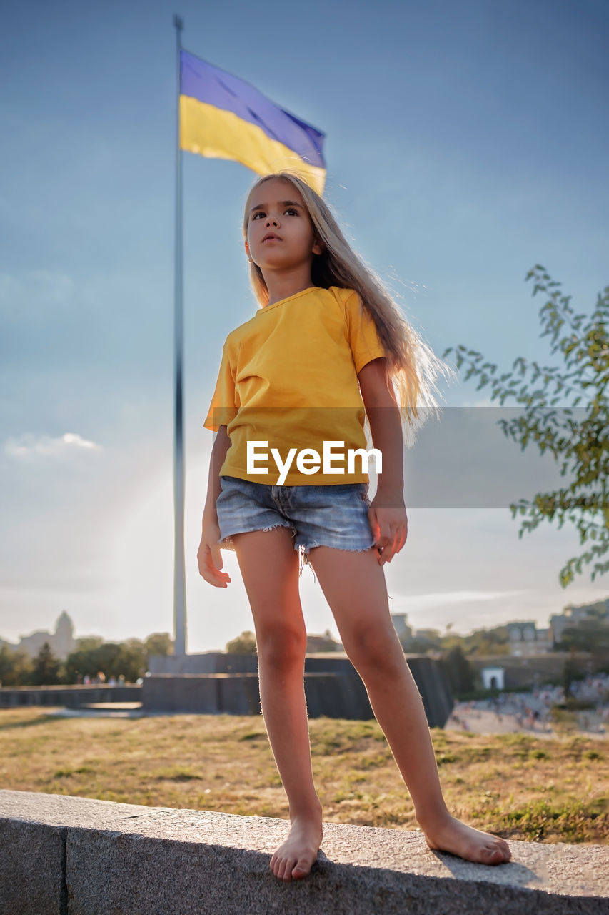 Girl in yellow polo and blue shorts near the national flag of ukraine. pray for peace and victory