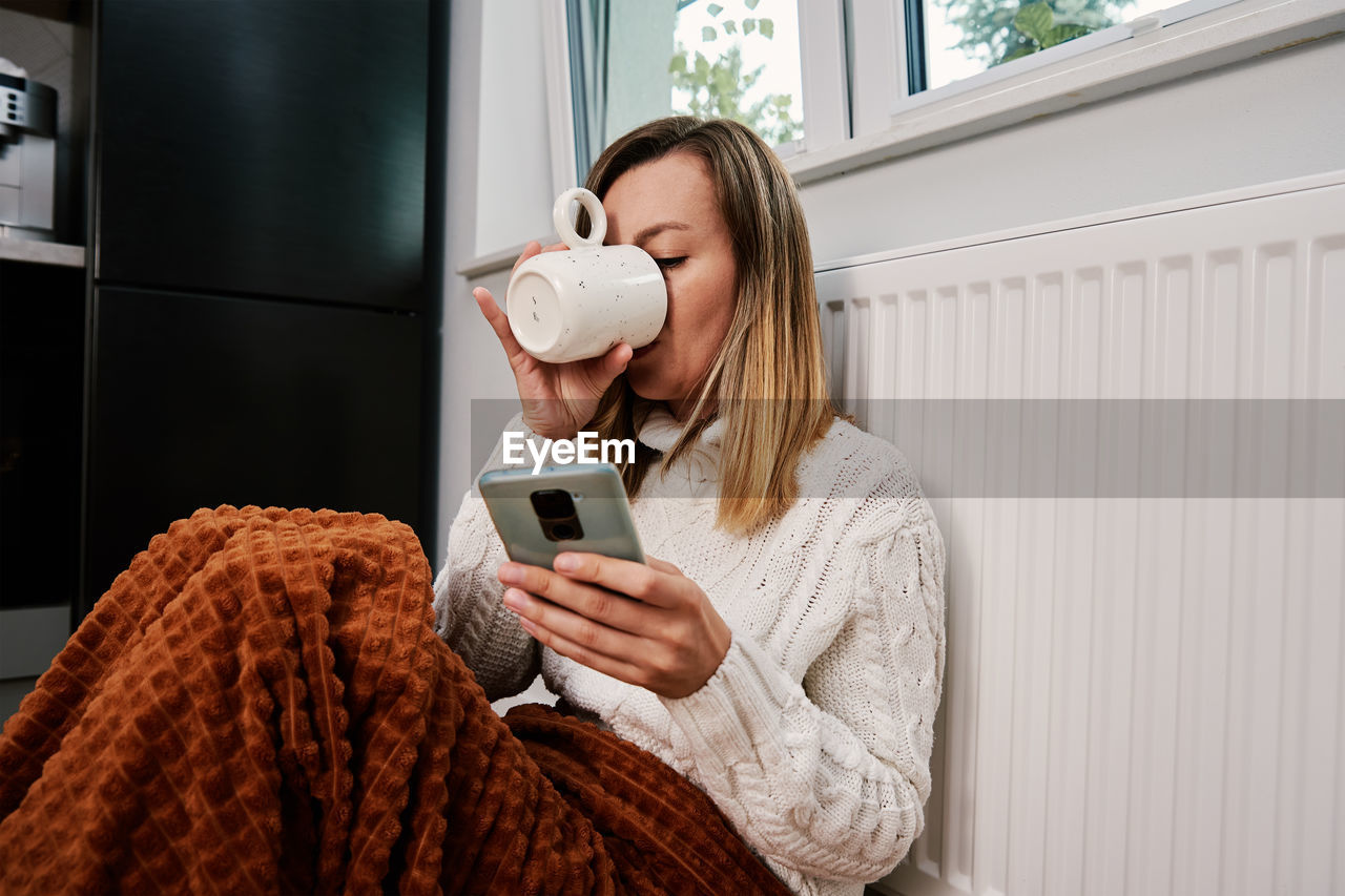 Worried woman sit near heating radiator under blanket and using smartphone