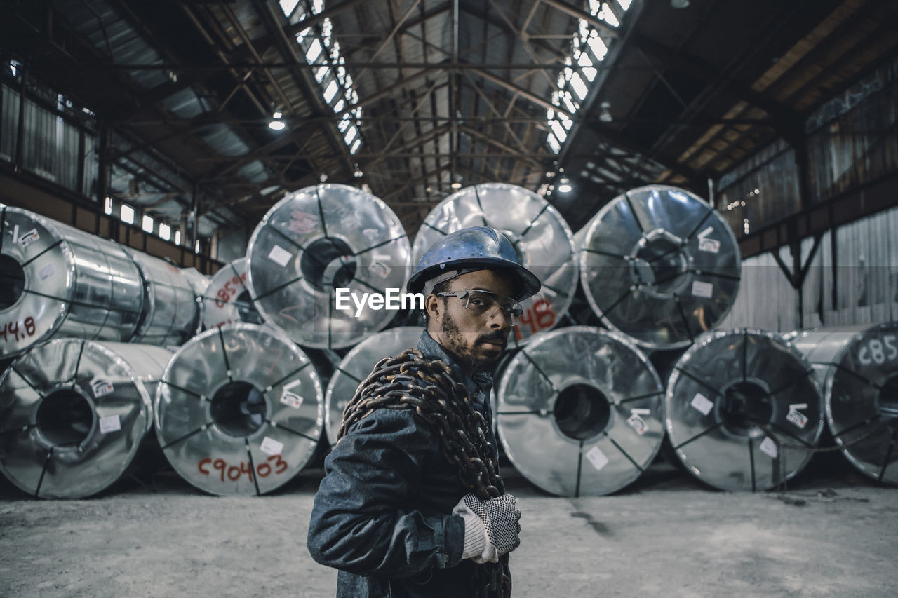 Portrait of male worker carrying chain while standing against rolled up steel sheets at factory