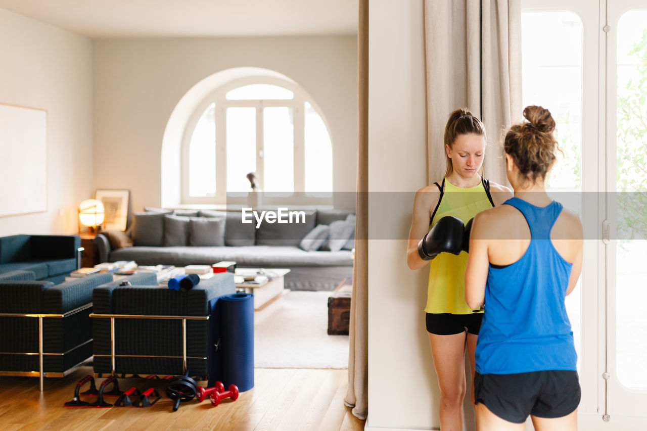 Serious athletic female in boxing gloves standing at home with personal instructor and preparing for workout