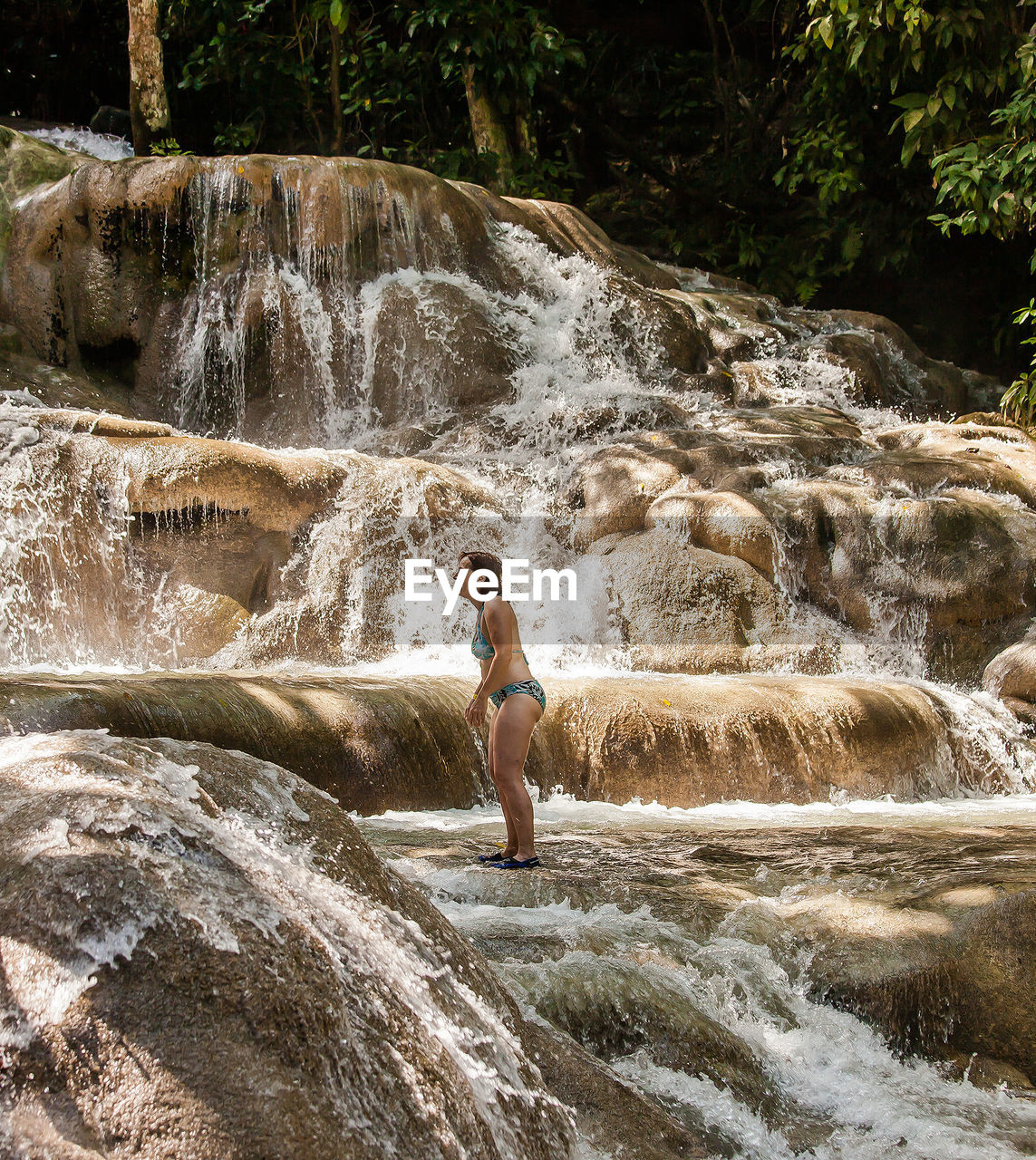 Side view of woman wearing bikini standing on rock against waterfall