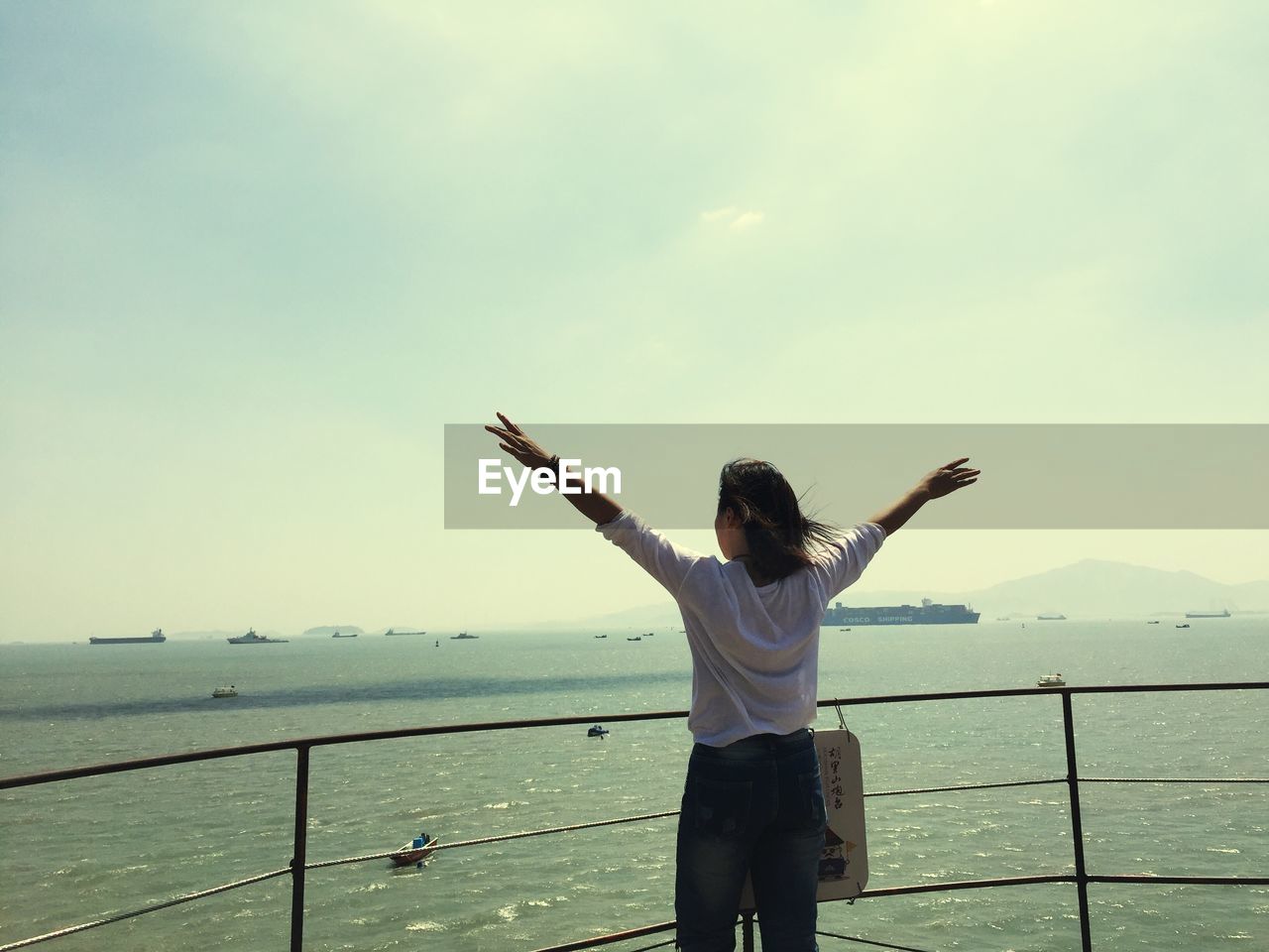 REAR VIEW OF MAN STANDING BY RAILING AGAINST SEA