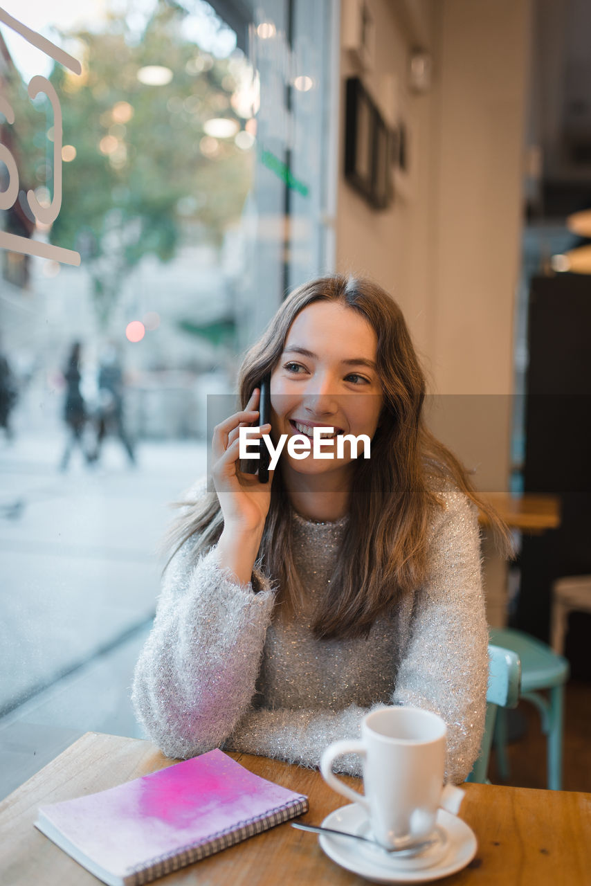 Content charming female sitting at table in cafe and speaking on smartphone while looking out of window and smiling