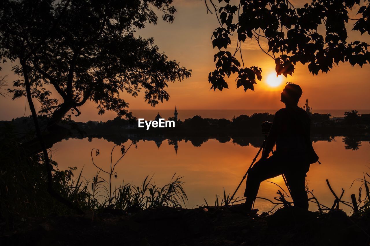 Silhouette mature man with camera standing by lake against sky during sunset