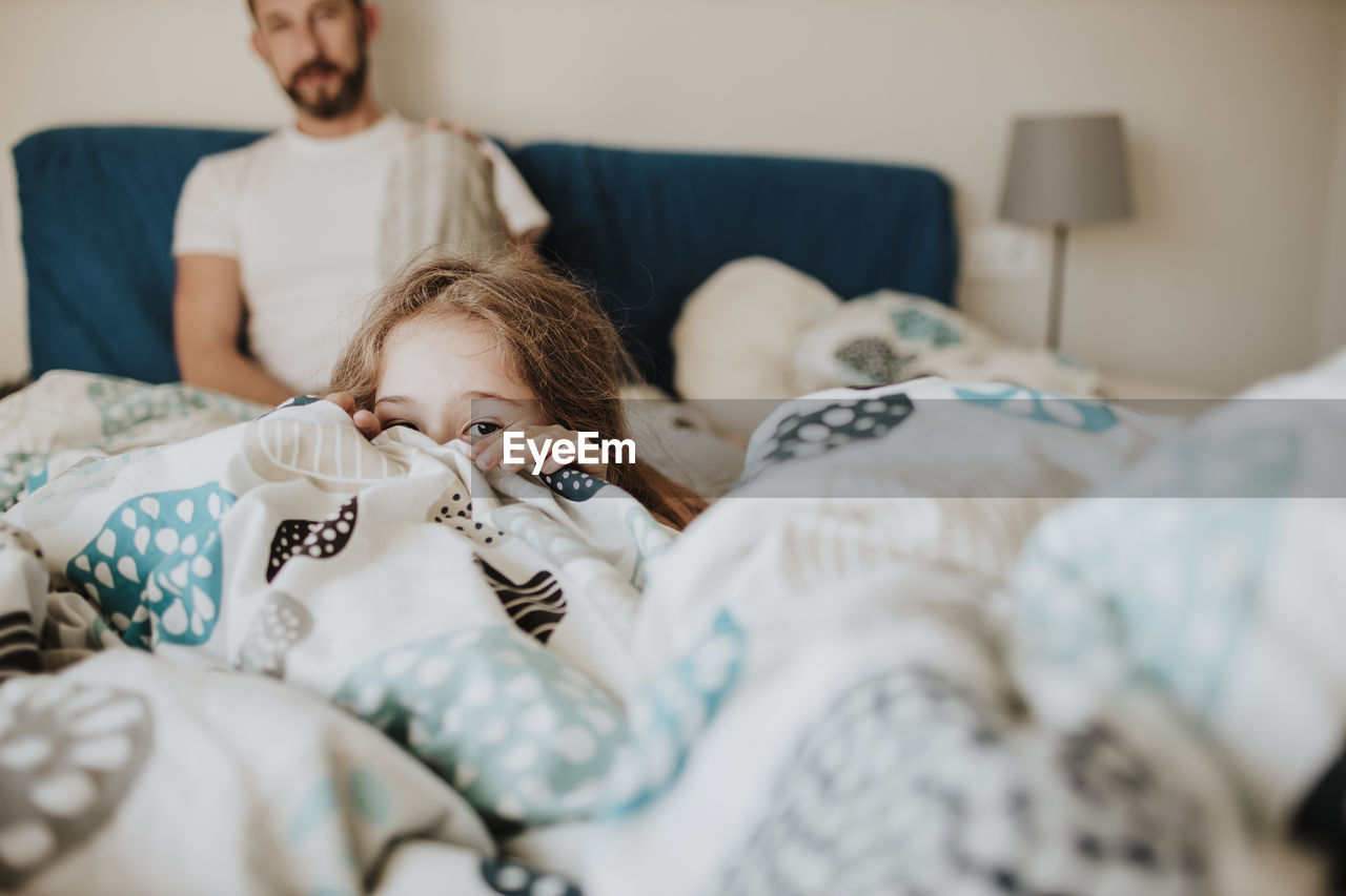 Girl hiding in blanket while father watching on bed at home