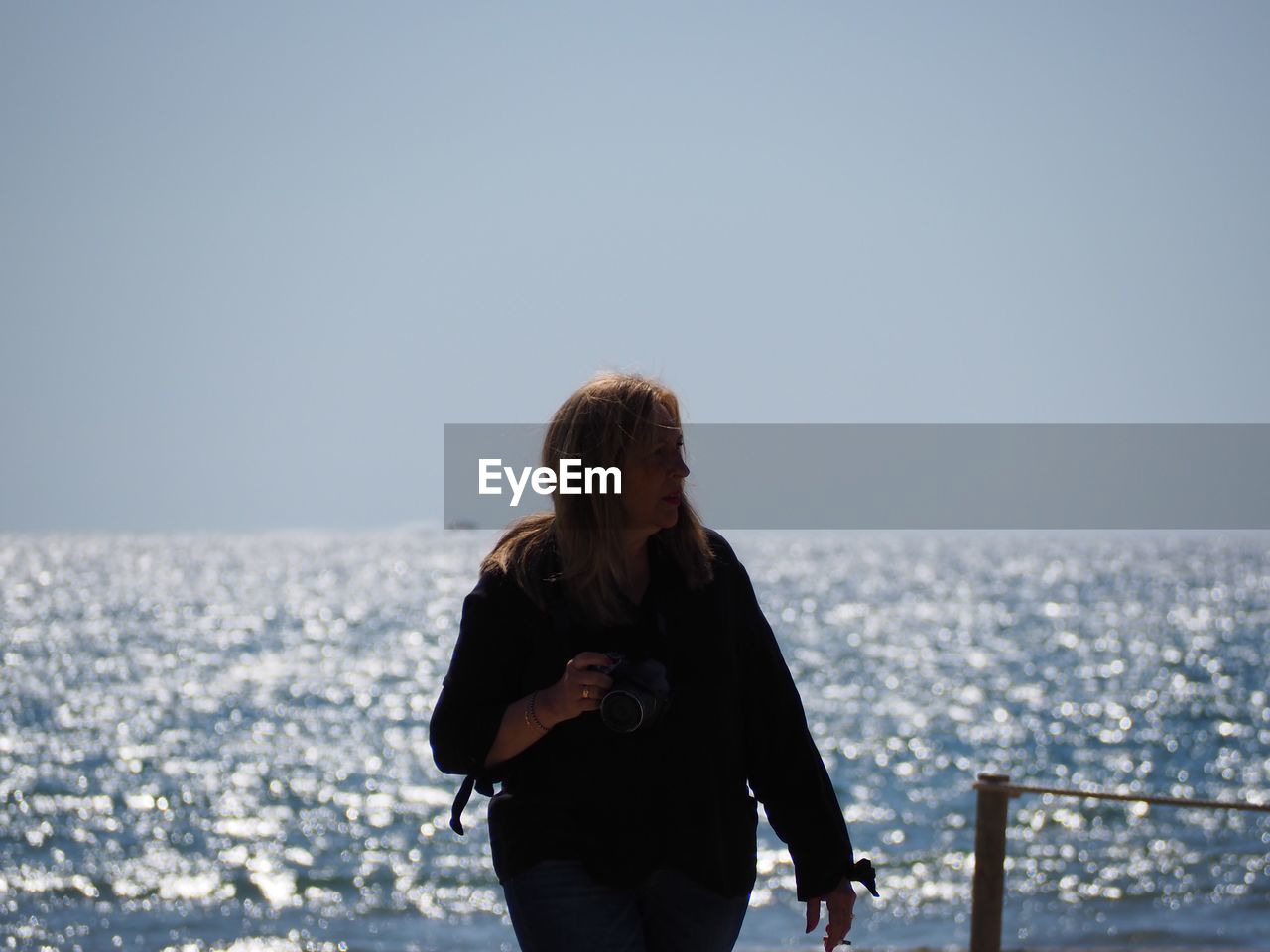 Mature woman holding camera while standing against sea at beach