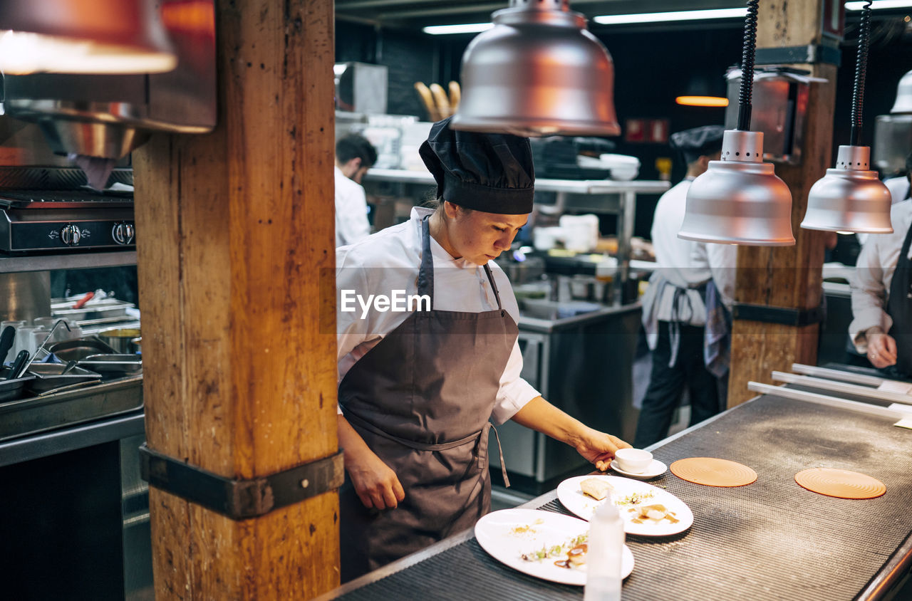 Adult woman in apron and hat preparing and serving dishes on kitchen in modern restaurant