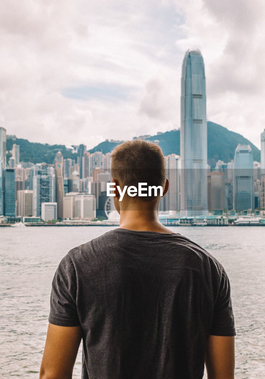 Rear view of young man standing by river in city against sky