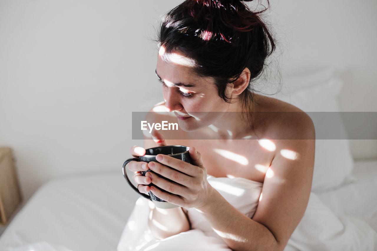 Woman sitting on bed while holding coffee cup at home