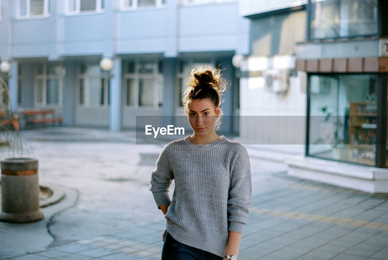 Portrait of young woman standing against building on street in city