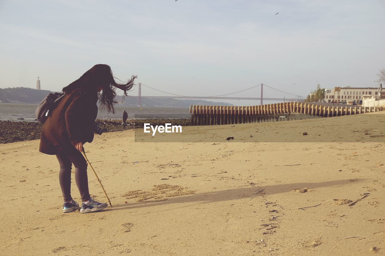 Full length of woman holding stick at sandy beach against sky