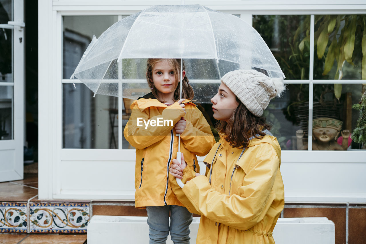 Sister looking at girl holding umbrella while standing on bench
