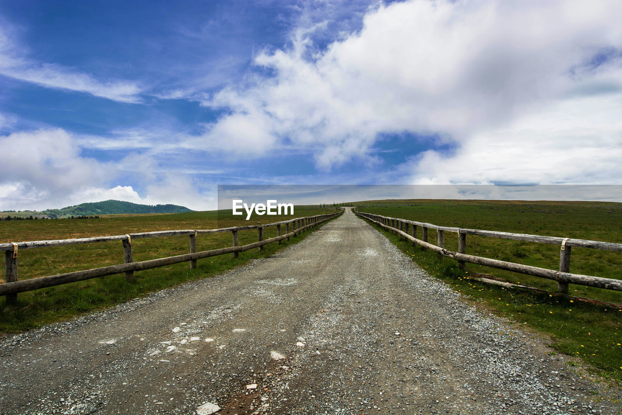 Road passing through field against cloudy sky