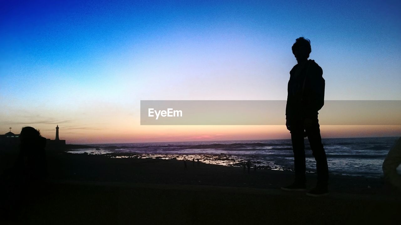 SILHOUETTE MAN STANDING ON BEACH AGAINST CLEAR SKY