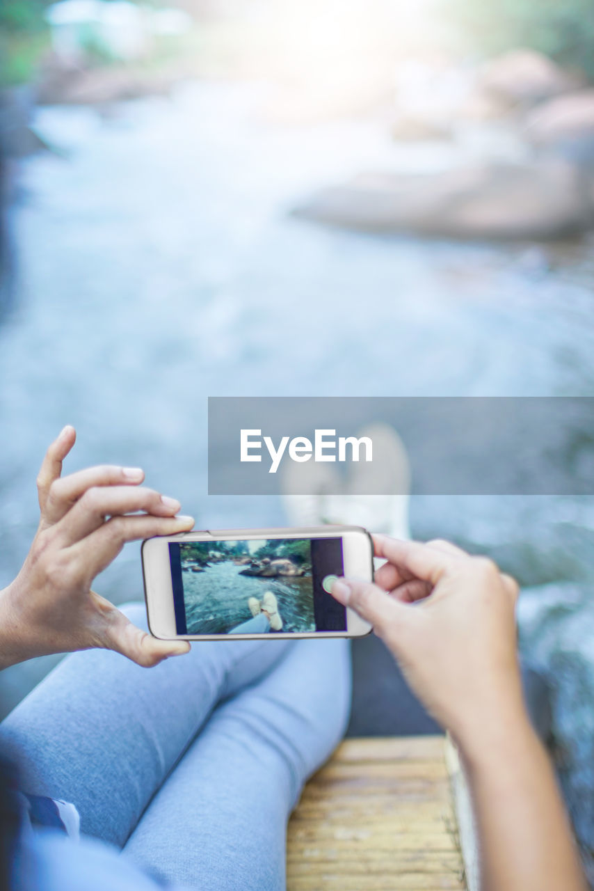 Low section of woman photographing with mobile phone while sitting by river