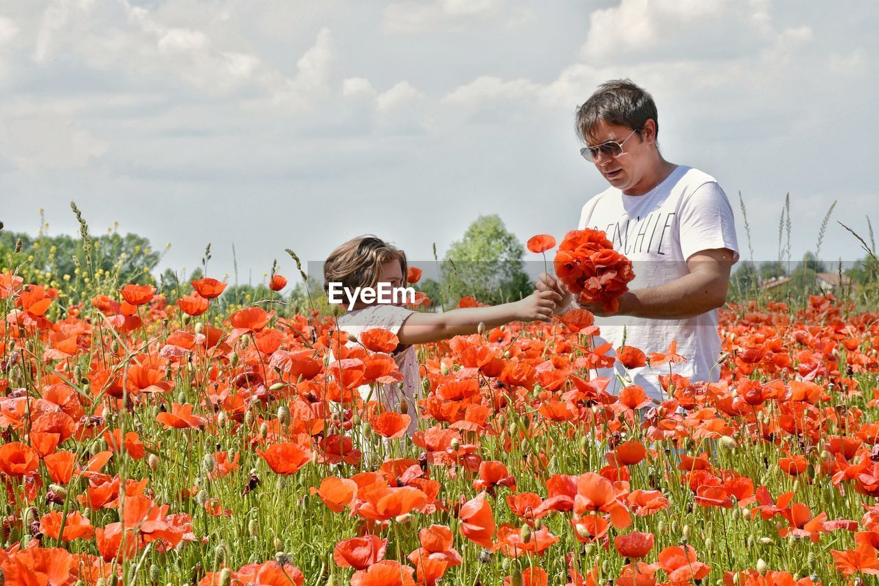 Father and daughter picking poppies from farm against cloudy sky