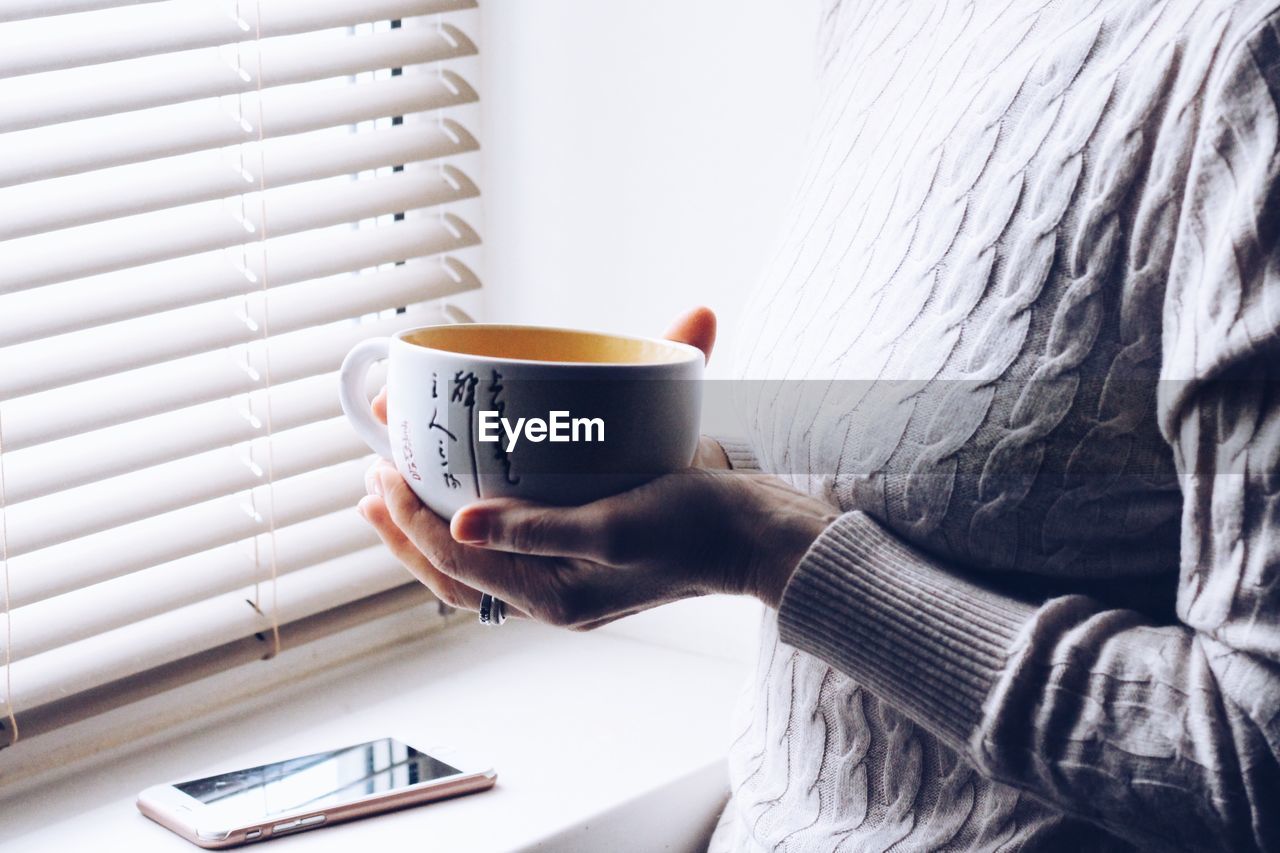 Midsection of woman holding coffee cup by window at home