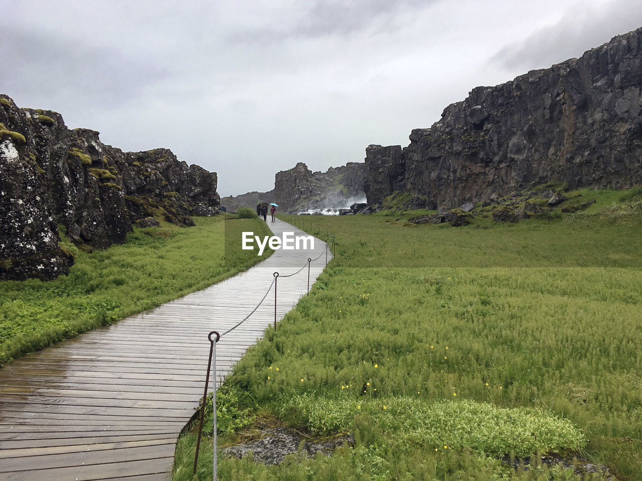 Boardwalk amidst grassy field and rock formations against cloudy sky