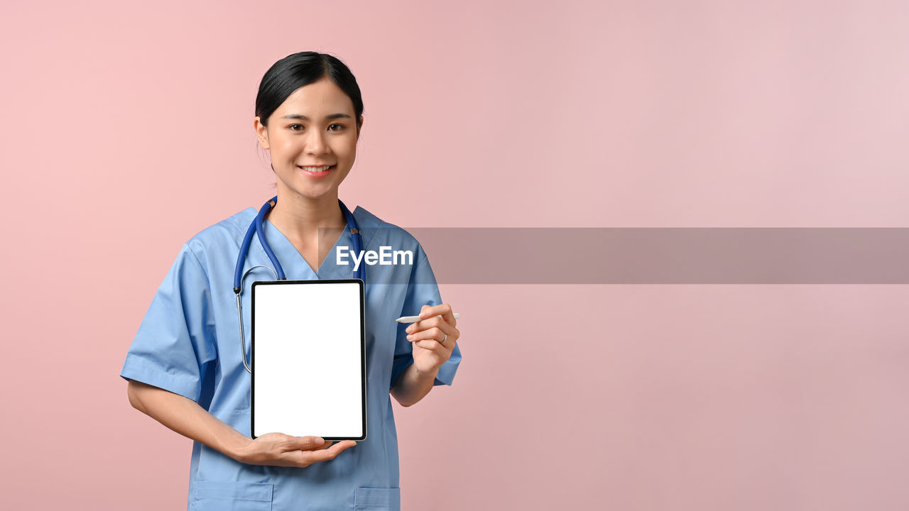 adult, technology, smiling, one person, digital tablet, copy space, portrait, looking at camera, women, standing, occupation, studio shot, colored background, indoors, communication, holding, computer, happiness, wireless technology, front view, waist up, female, cheerful, doctor, working, young adult, computer network, business, internet, medical instrument, stethoscope, professional occupation, medical equipment, healthcare and medicine, showing, using computer, emotion, healthcare worker, expertise, black hair, portability, three quarter length