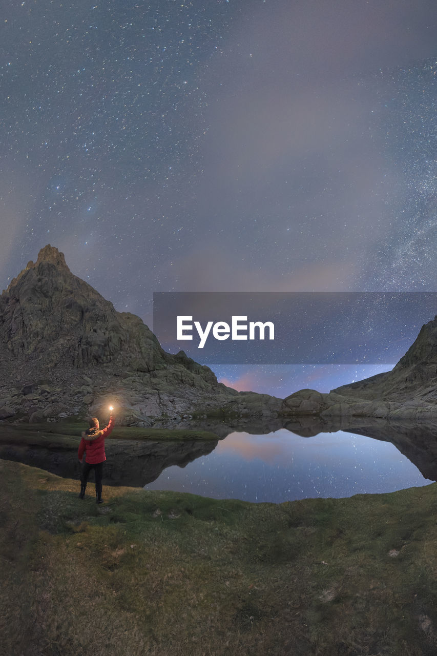 Back view of unrecognizable male tourist with torch admiring snowy mounts reflecting in water under starry sky at night