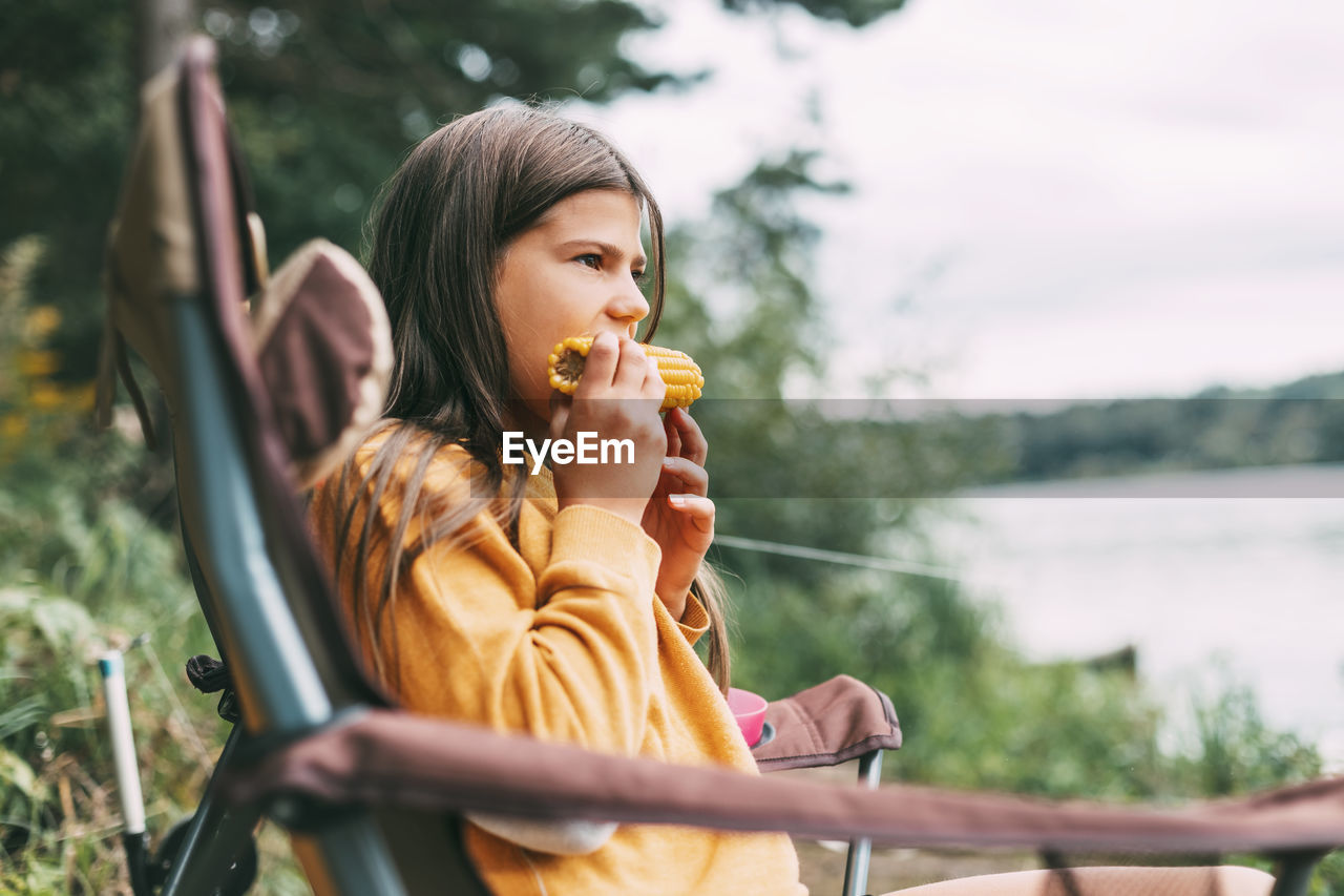 A teenage girl in a bright yellow sweater is sitting in a camping chair on the shore of the lake 