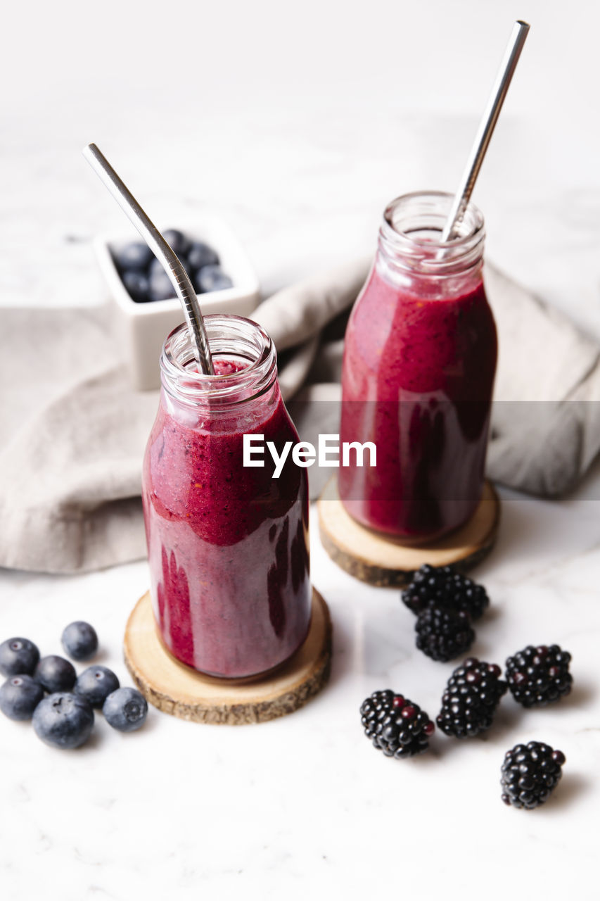 Berry smoothie and metal straws on glass bottles on wood slices over a white marble surface. high angle, vertical image.