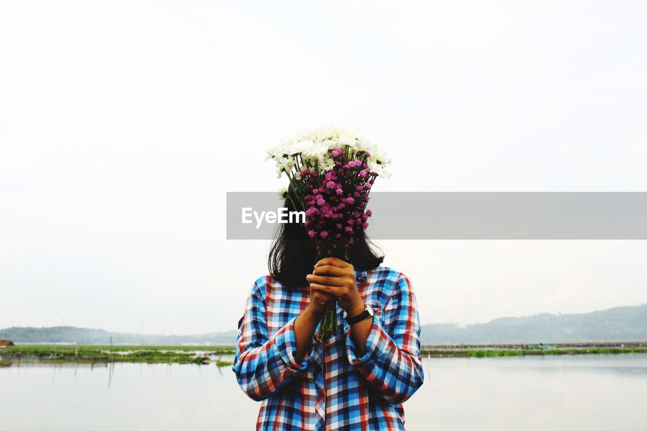 Woman covering face with bouquet by lake against clear sky