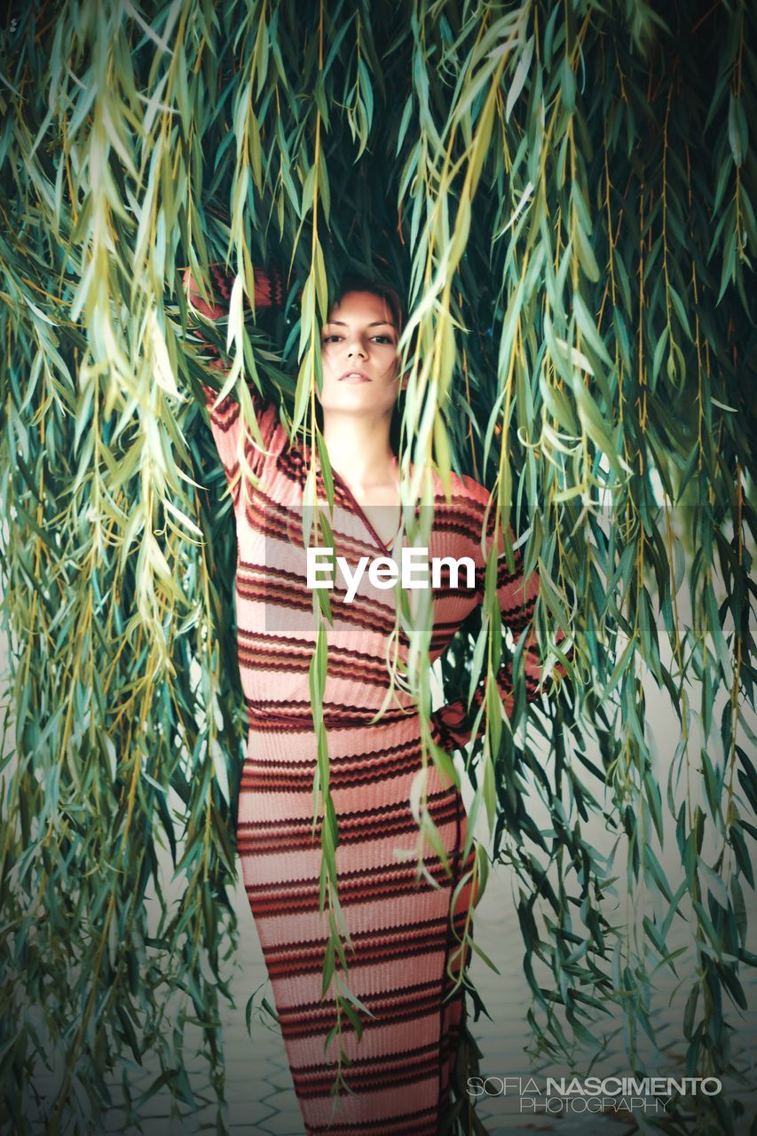 PORTRAIT OF SMILING YOUNG WOMAN STANDING BY PLANTS