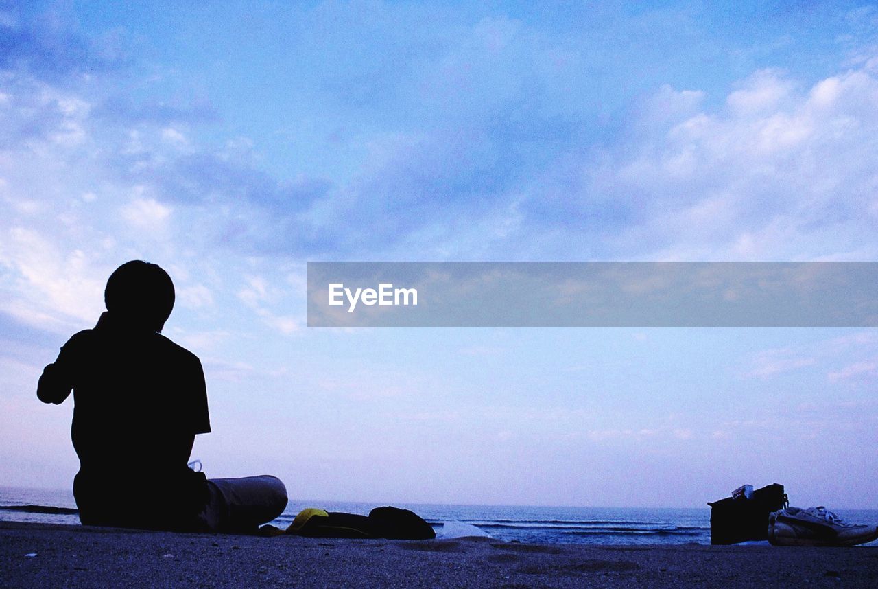 SILHOUETTE BOY SITTING ON SHORE AGAINST SKY