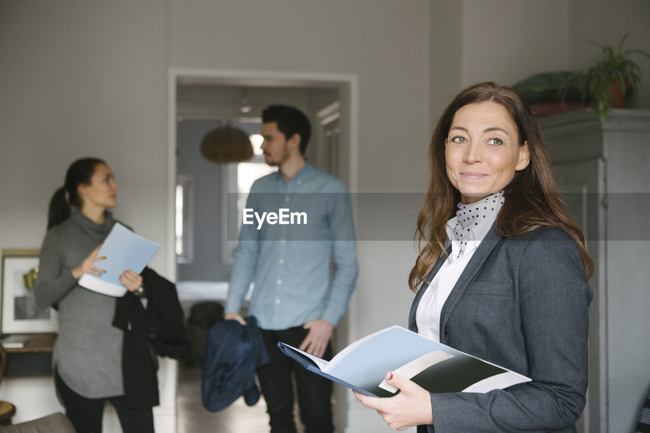 Confident female realtor holding brochure while couple standing in background