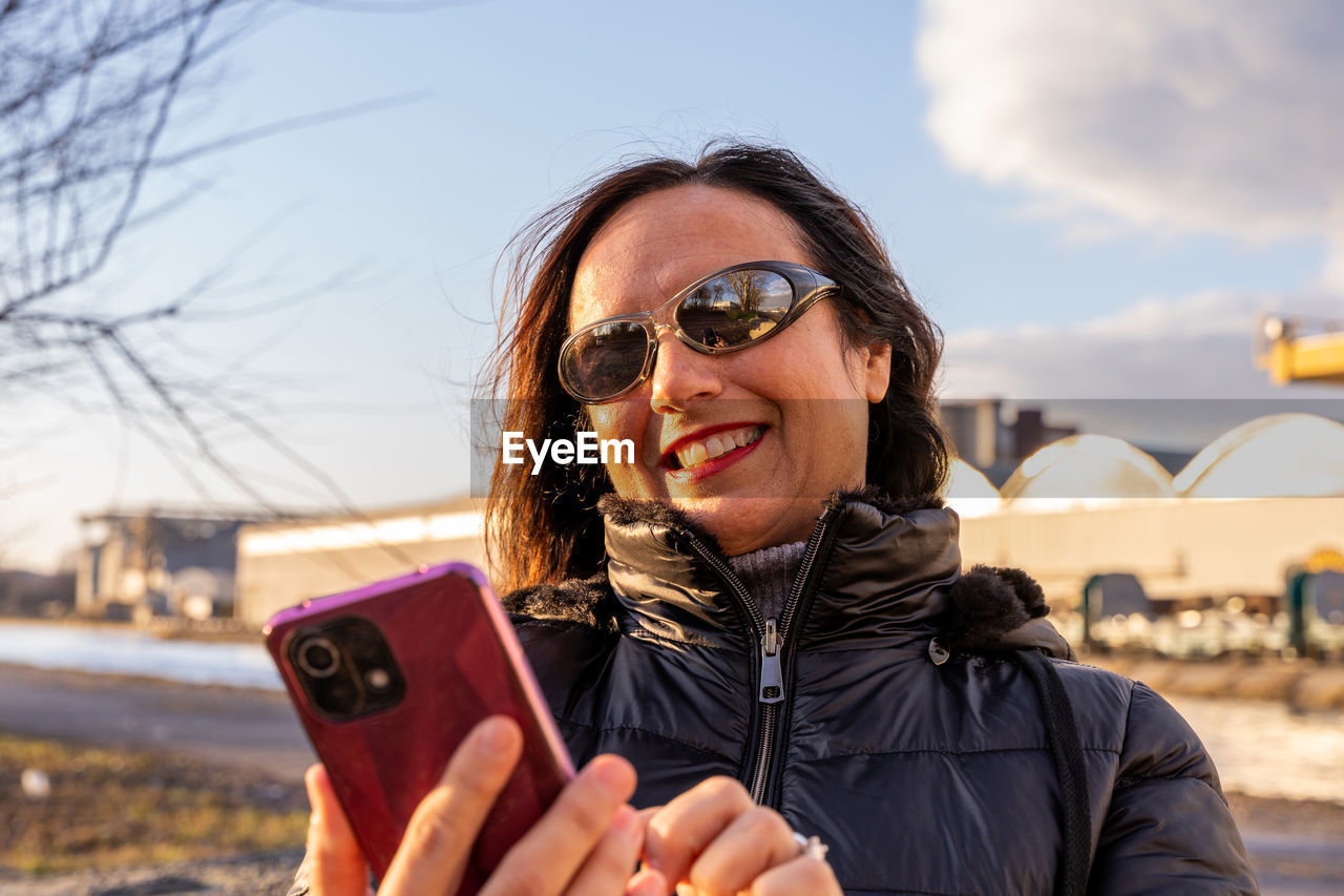 Middle aged woman wearing winter clothes using smartphone in front of a factory - people having fun