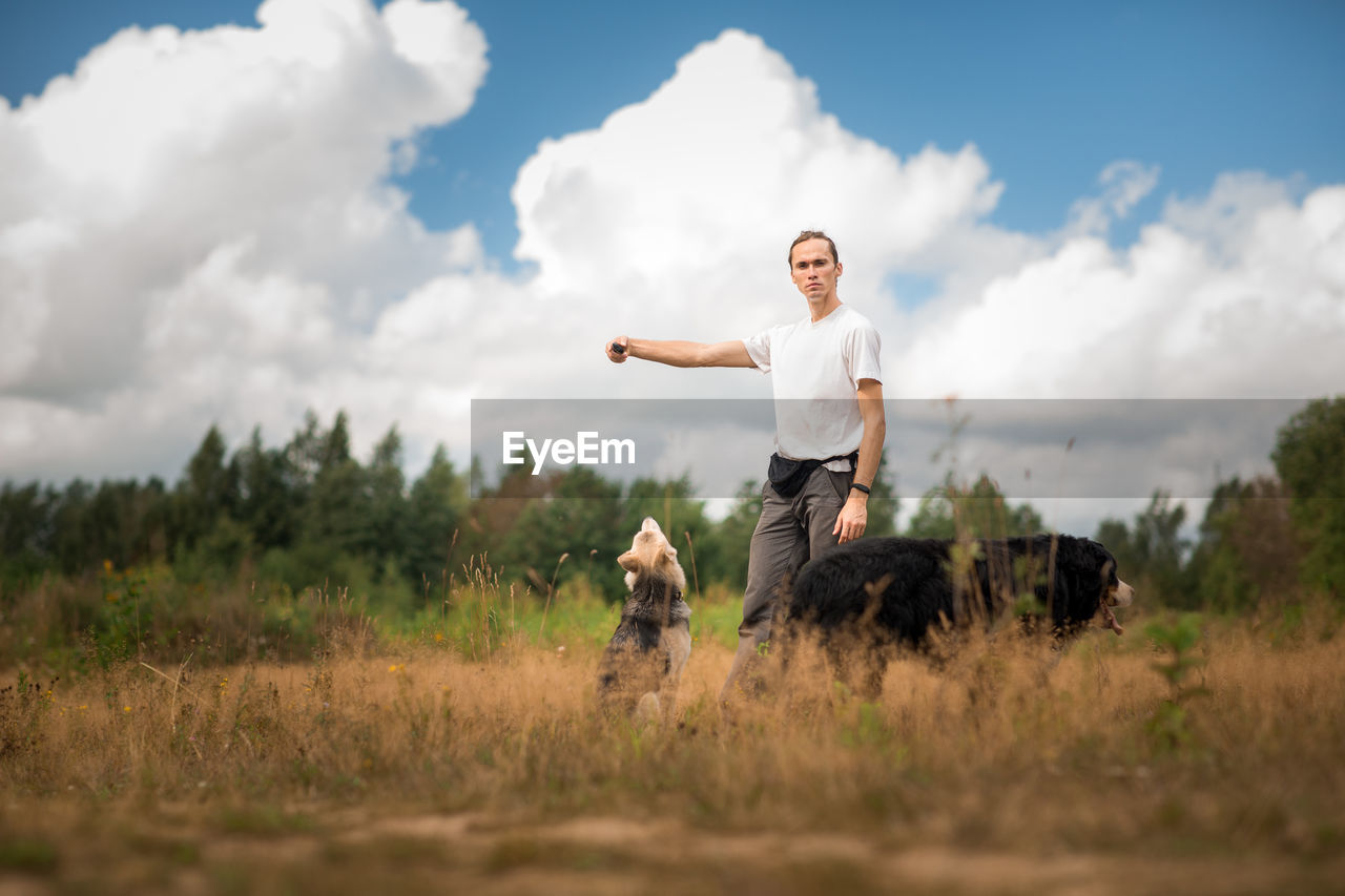 Portrait of young man with dogs on grassy land against sky