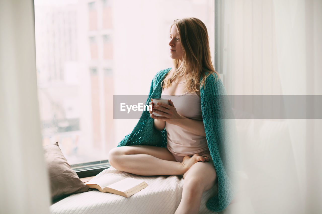 A young girl is sitting on the windowsill in the early morning, drinking hot coffee