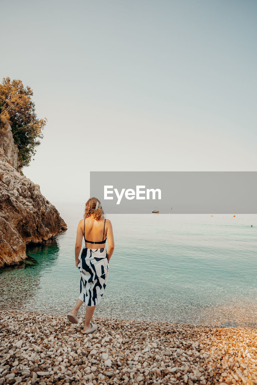water, sea, land, beach, sky, nature, shore, one person, adult, full length, coast, sand, horizon, copy space, beauty in nature, ocean, rock, holiday, vacation, leisure activity, trip, tranquility, body of water, women, lifestyles, young adult, day, scenics - nature, clothing, travel, clear sky, tranquil scene, outdoors, relaxation, rear view, morning, sunlight, standing, travel destinations, horizon over water, summer, casual clothing, idyllic, coastline, solitude, person, environment, sunny, emotion, wave, motion