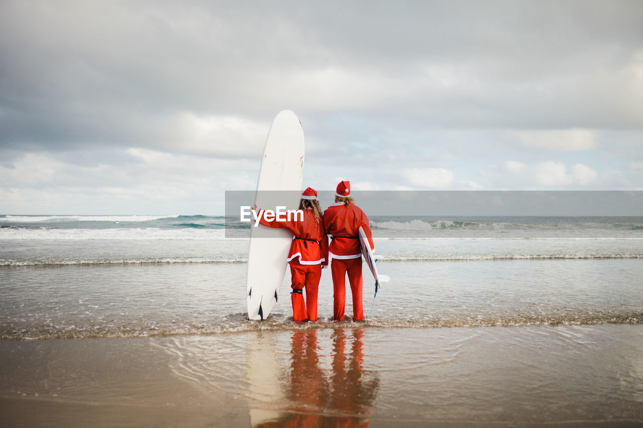 Rear view of couple wearing santa costumes holding surfboards in sea