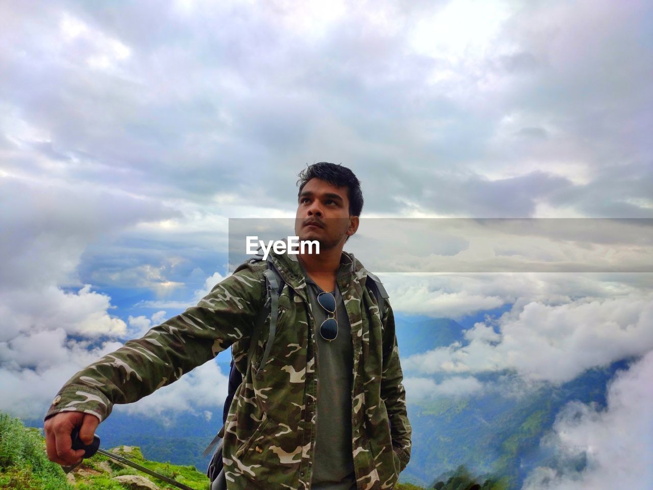 Young man looking away while standing on mountain against cloudy sky