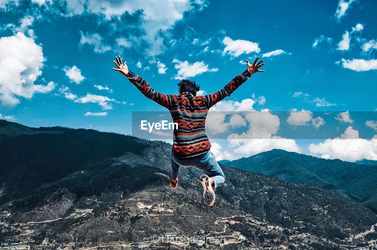 Rear view of playful man jumping with mountains in background against blue sky