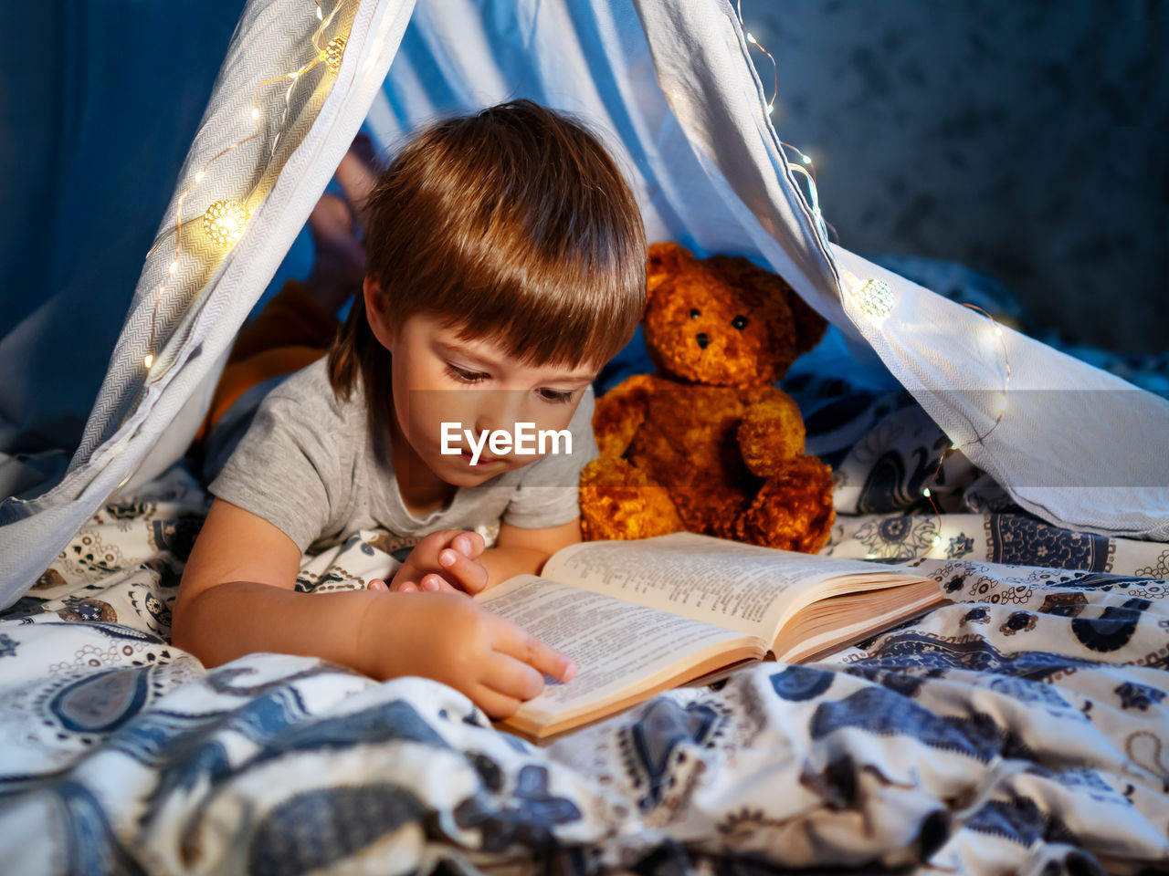 Little boy reads book with teddy bear. toddler plays in tent made of linen sheet on bed. coziness.