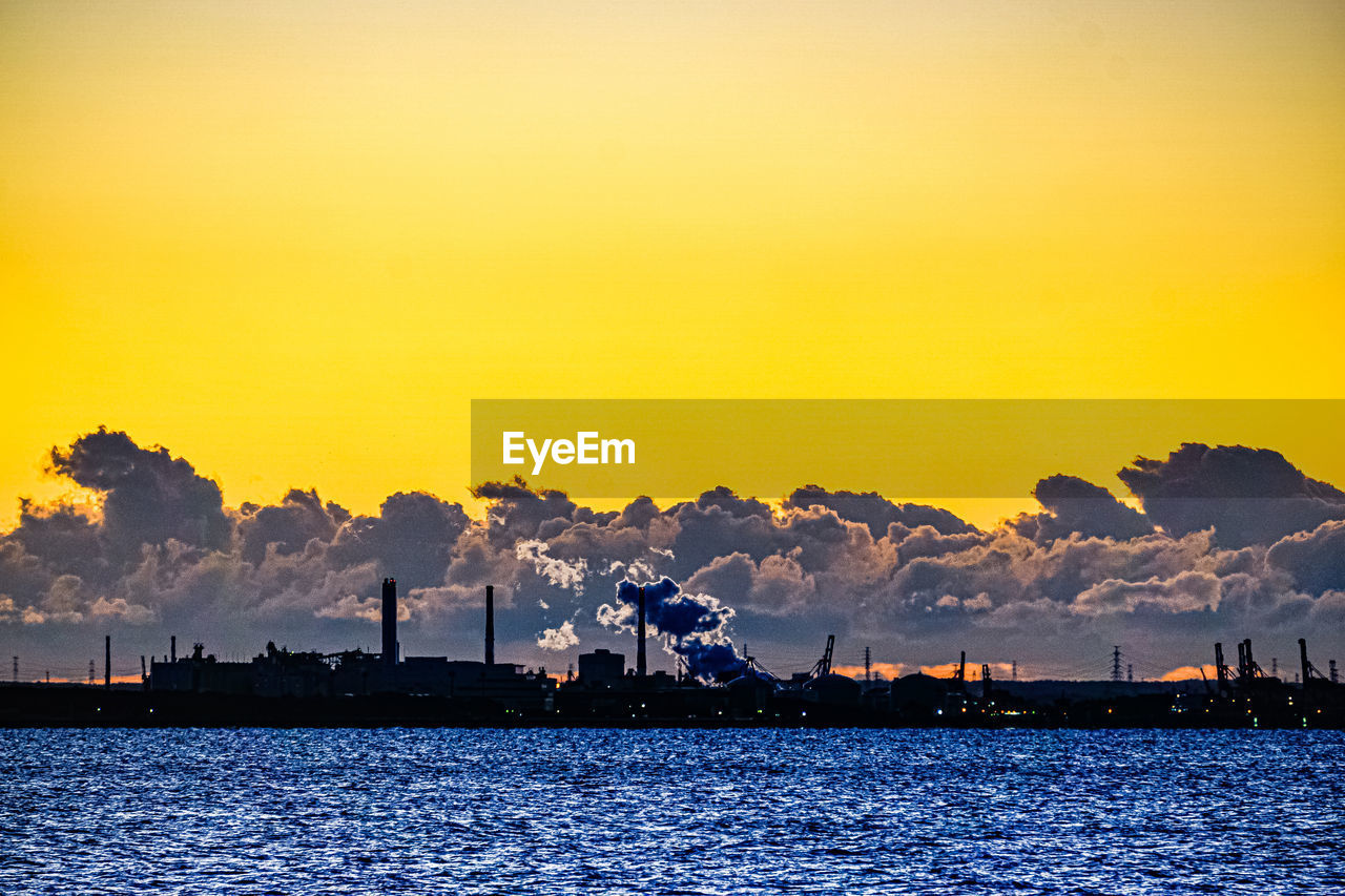 SILHOUETTE OF FACTORY BY SEA AGAINST SKY