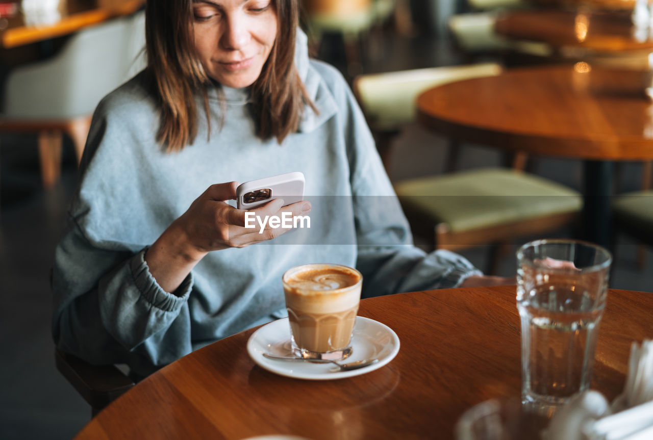 Young brunette woman wearing casual longsleeve with mobile phone in hands with cup of coffee in cafe