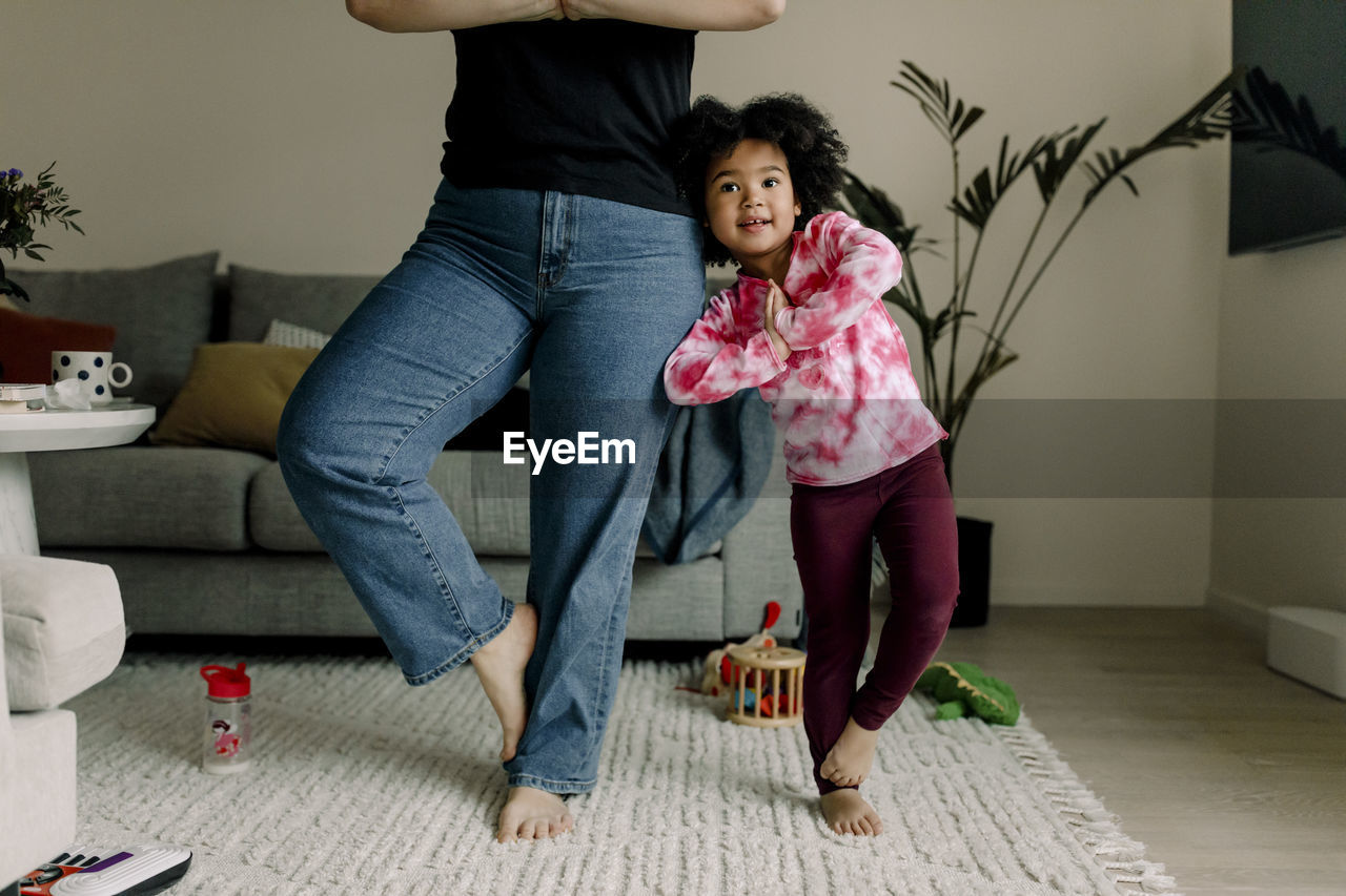 Mother and daughter doing tree pose while practicing yoga in living room