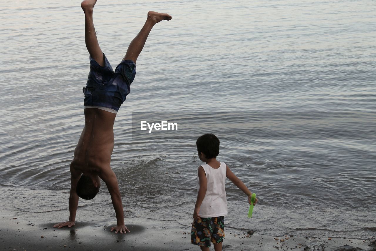 Boy looking at brother doing handstand on sea shore at beach