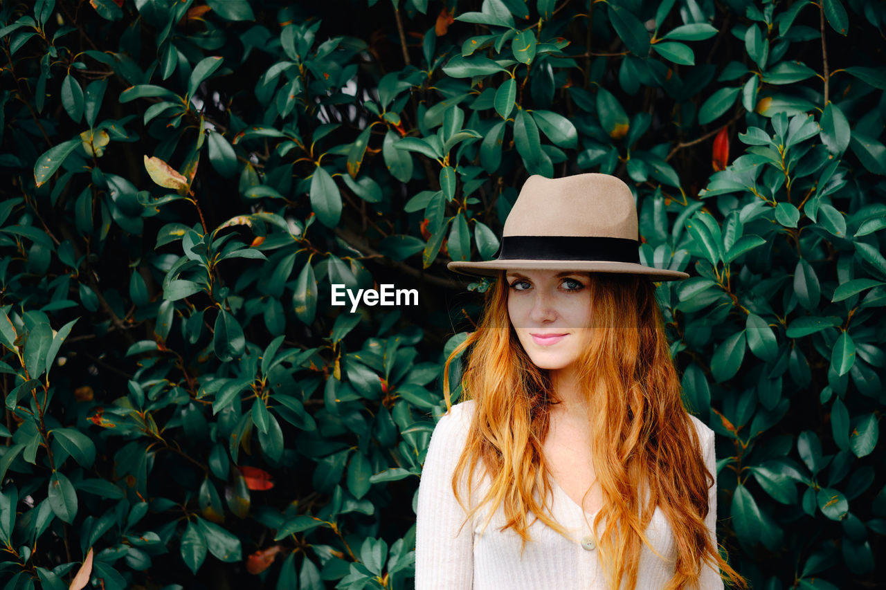 Attractive young long haired ginger female in stylish hat looking at camera while standing against green bushes in summer garden