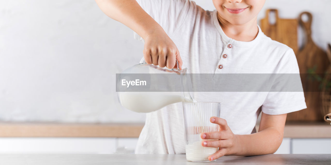 Cute smiling boy pours fresh milk from a jug into a mug in the kitchen.