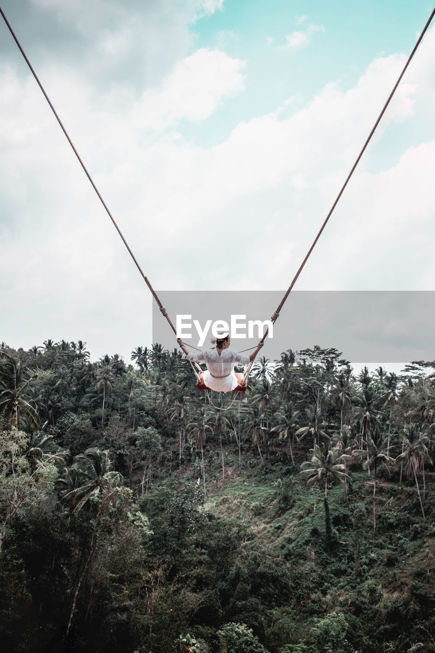 Woman sitting on swing by palm trees against sky