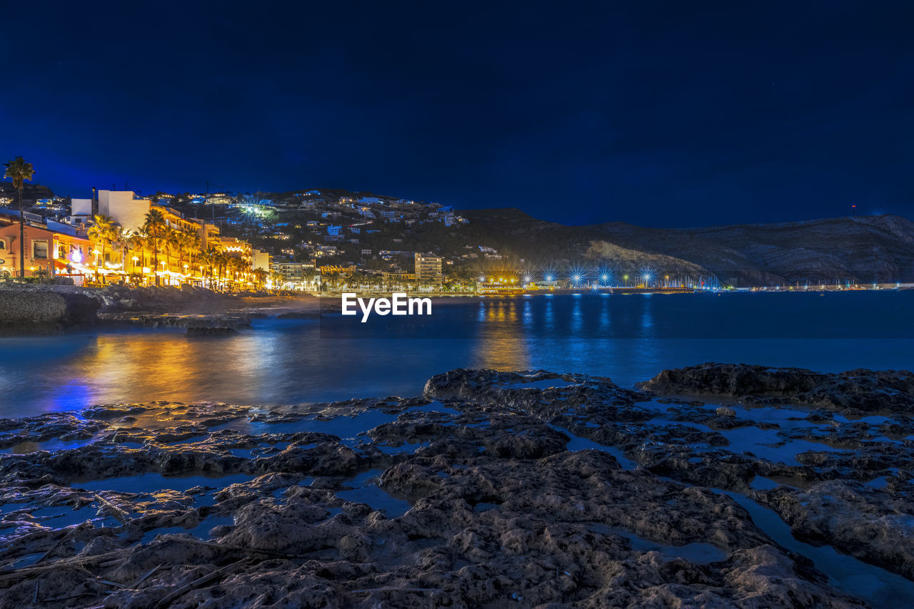 Illuminated buildings by sea against sky at night