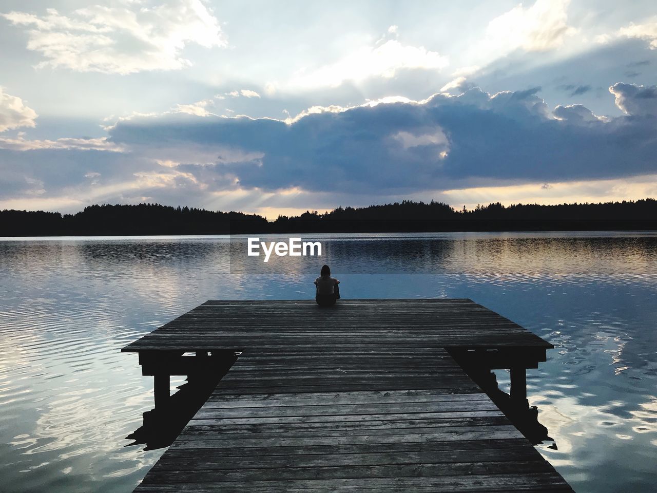 Woman sitting on pier over lake against sky during sunset